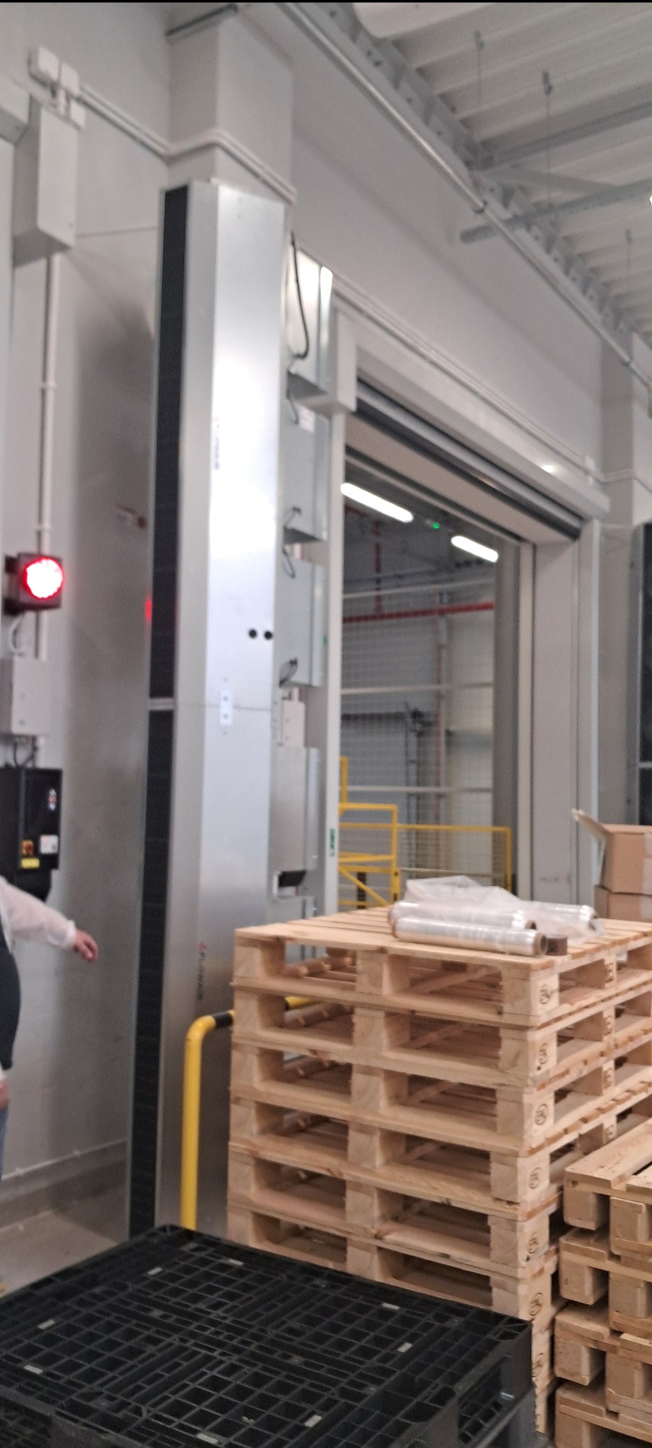 Wysoka, metalowa kolumna wentylacyjna w hali magazynowej, z widocznymi elementami instalacji i paletami w tle. Oświetlenie sufitowe i brama wjazdowa.