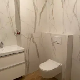 Wykończona łazienka z białymi płytkami imitującymi marmur, wiszącą toaletą i szafką z umywalką. Podłoga wyłożona panelami imitującymi drewno. Nowoczesny design.