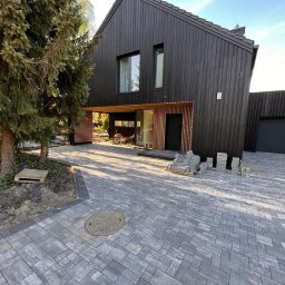 Aurora Prime Sp. z o.o. - Nowoczesny dom z czarną elewacją i szarą kostką brukową na podjeździe. Widoczne palety z kostką, sugerujące trwające prace wykończeniowe.