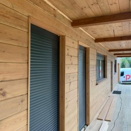 Aurora Prime Sp. z o.o. - Drewniana elewacja budynku z zamontowanymi, opuszczonymi roletami zewnętrznymi w kolorze grafitowym. Widok z zewnątrz, z boku budynku.