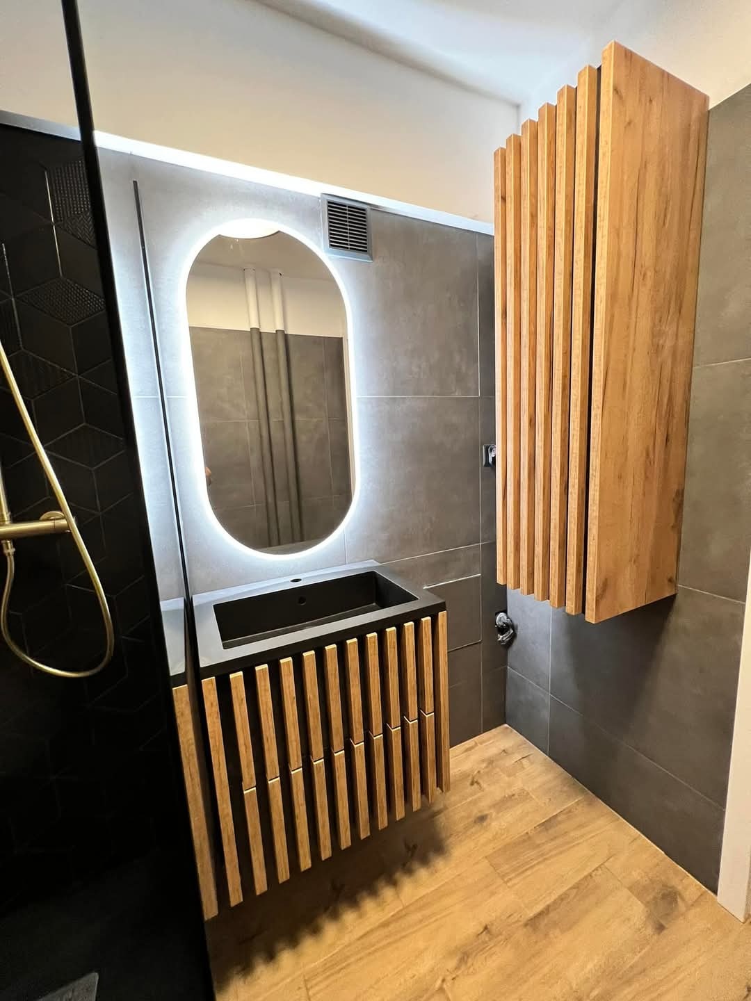 Nowoczesna łazienka z drewnianymi akcentami: szafka pod umywalkę i wisząca szafka z lamelami, podświetlane lustro owalne, szare płytki i drewnopodobna podłoga.