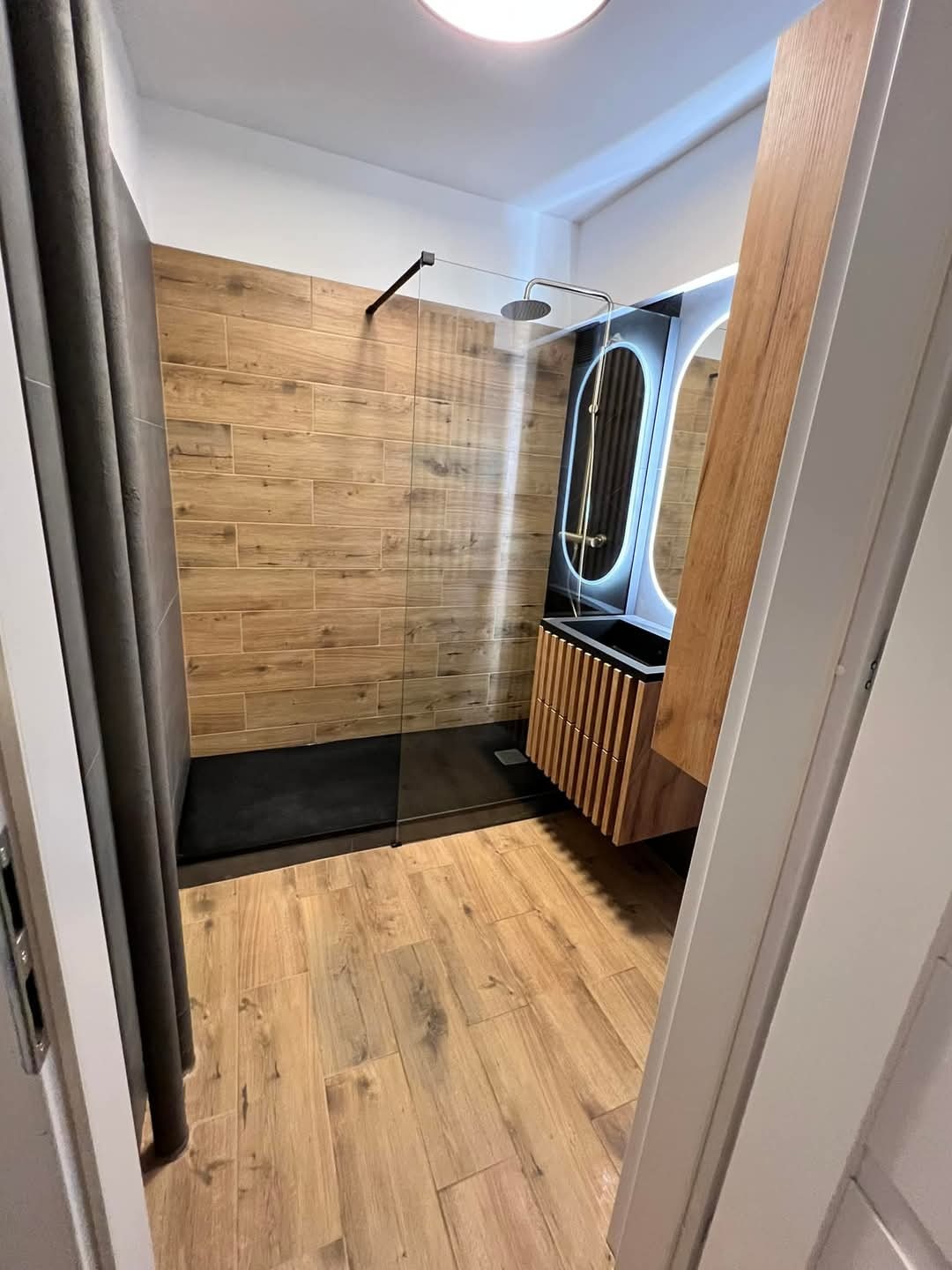 Nowoczesna łazienka z drewnopodobnymi płytkami, kabiną walk-in i podświetlanym lustrem. Ujęcie zza drzwi, widoczna armatura i szafka z lamelami.