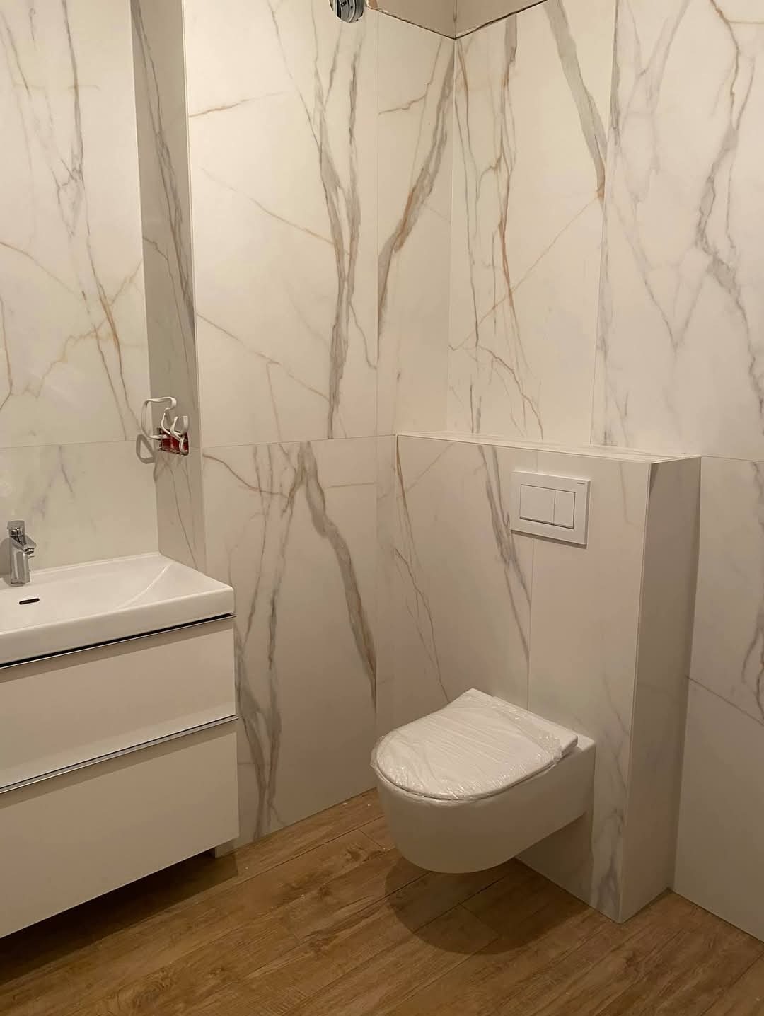 Wykończona łazienka z białymi płytkami imitującymi marmur, wiszącą toaletą i szafką z umywalką. Podłoga wyłożona panelami imitującymi drewno. Nowoczesny design.