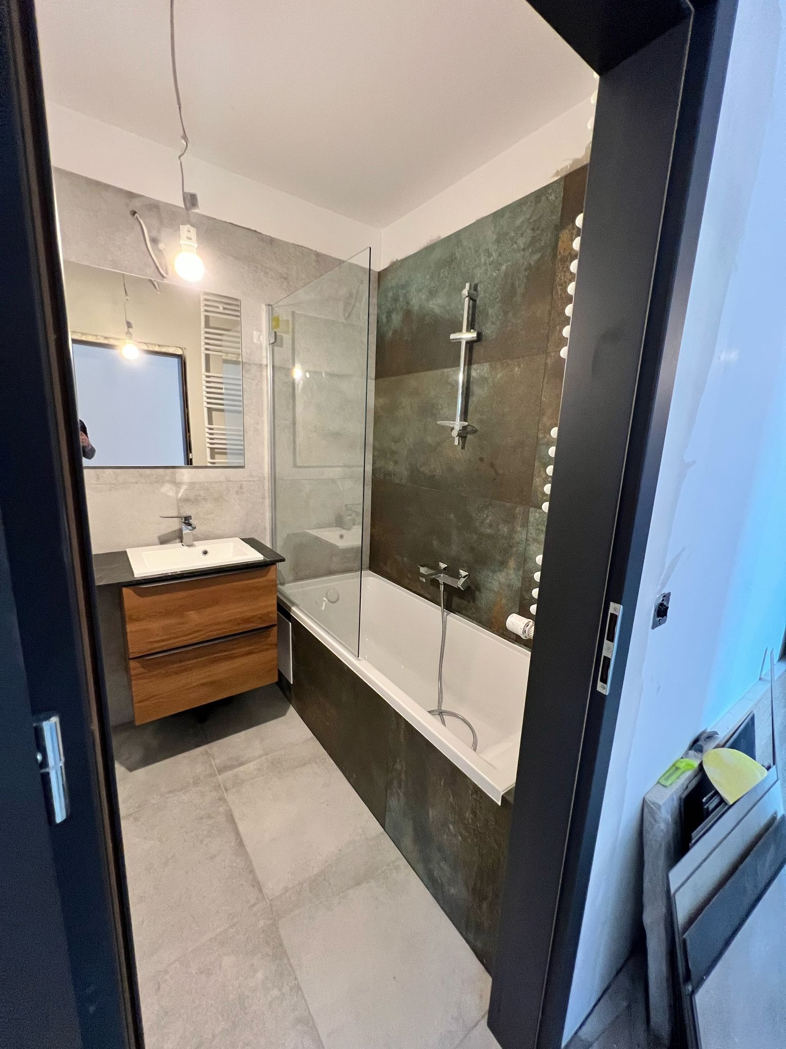 Nowoczesna łazienka z wanną, umywalką z drewnianą szafką i szklanym parawanem. Ciemne płytki imitujące rdzę kontrastują z jasnymi elementami wyposażenia.