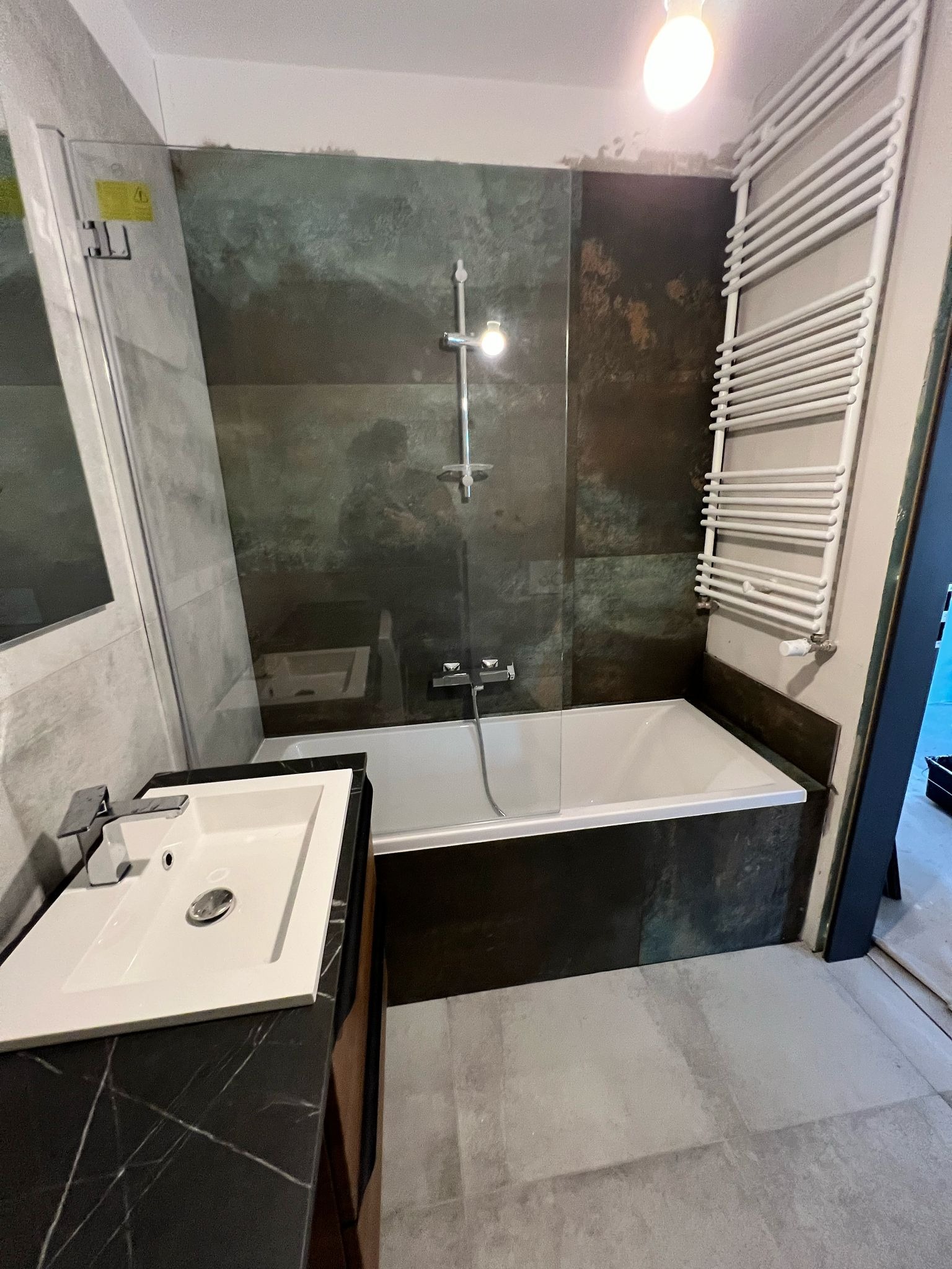 Wykończona łazienka z wanną za szklaną ścianą i umywalką na blacie z czarnego marmuru. Na ścianie grzejnik drabinkowy. Widoczne szare płytki podłogowe i dekoracyjne na ścianach.