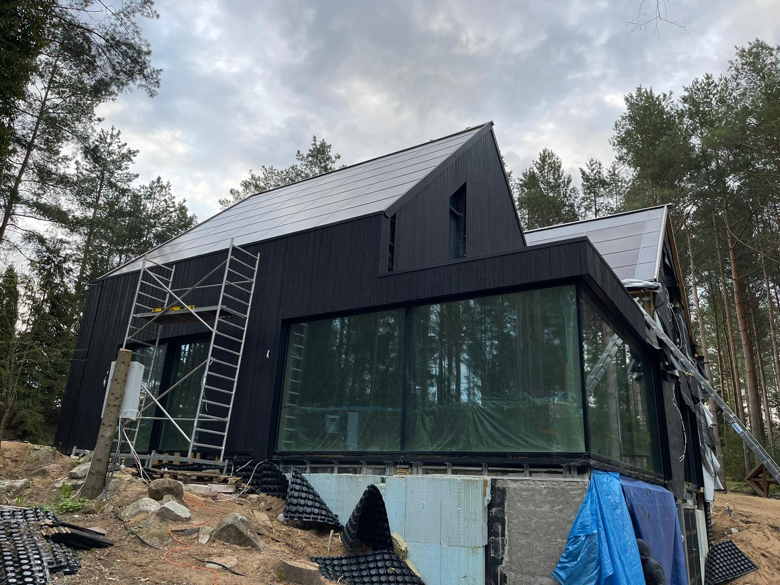 Czarny, nowoczesny dom w lesie w trakcie budowy, z widocznymi rusztowaniami, dużymi oknami i panelami na dachu. Otoczenie leśne.