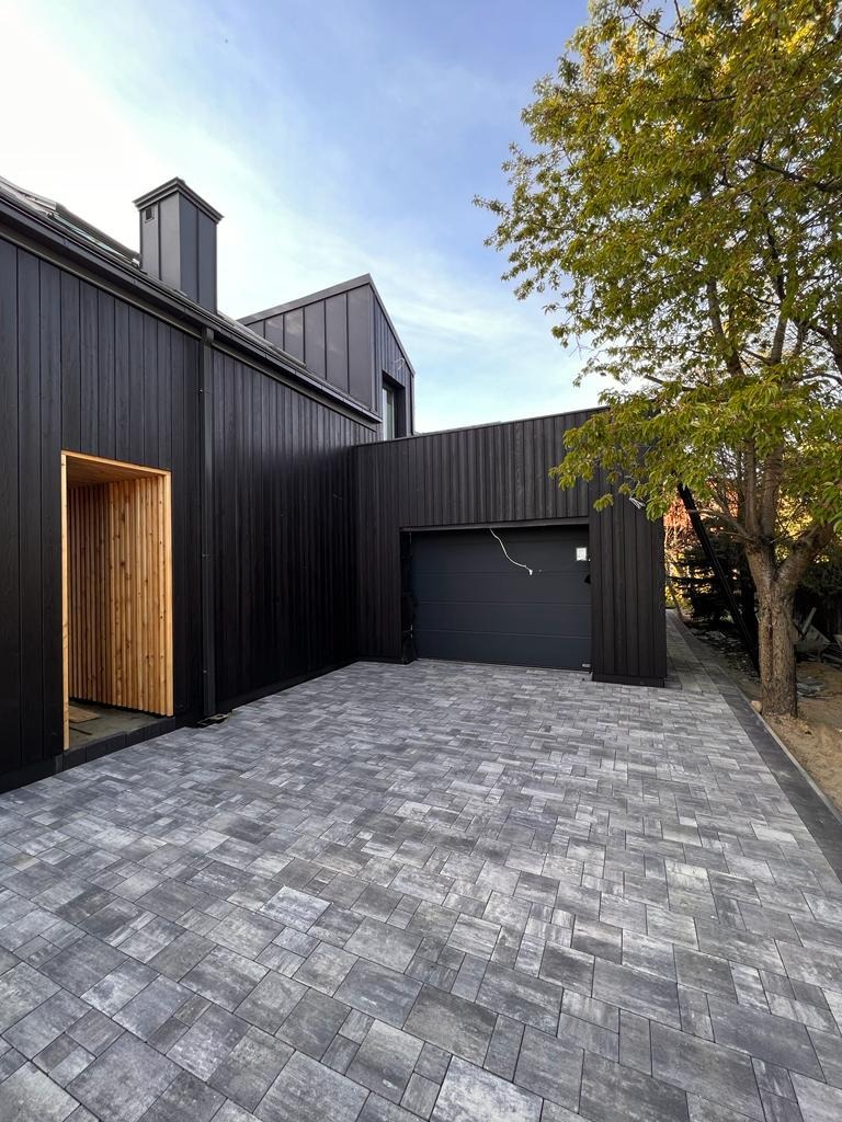 Nowoczesny dom z ciemną, drewnianą elewacją, szarą kostką brukową i garażem. Widoczny fragment drzewa i błękitne niebo w tle.