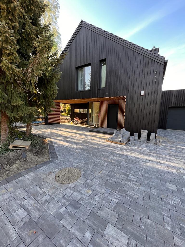 Nowoczesny dom z czarną elewacją i szarą kostką brukową na podjeździe. Widoczne palety z kostką, sugerujące trwające prace wykończeniowe.