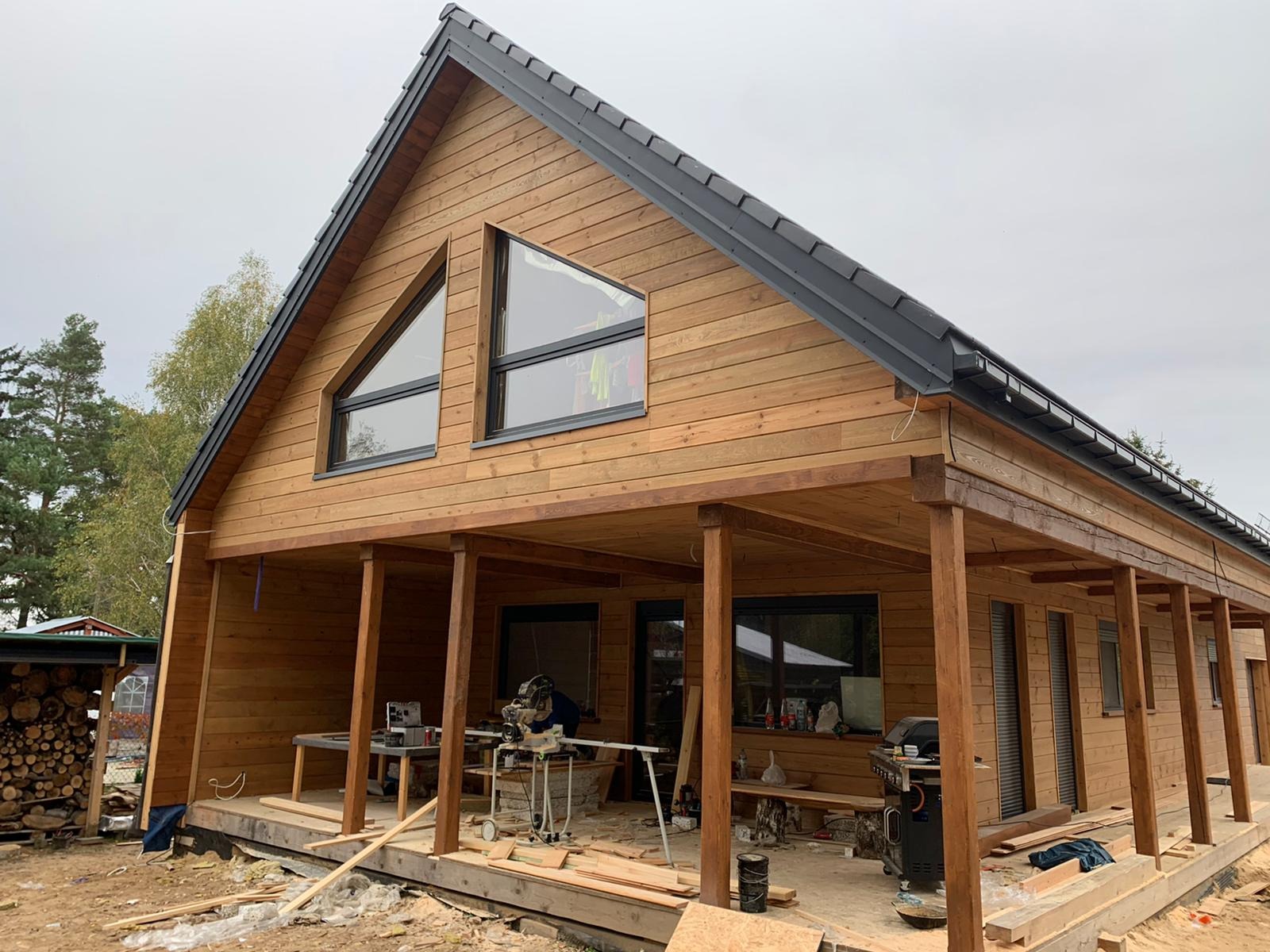 Drewniany dom z czarną dachówką i oknami o nietypowym kształcie. Częściowo zadaszony taras z widocznymi narzędziami i materiałami budowlanymi. Prace wykończeniowe w toku.