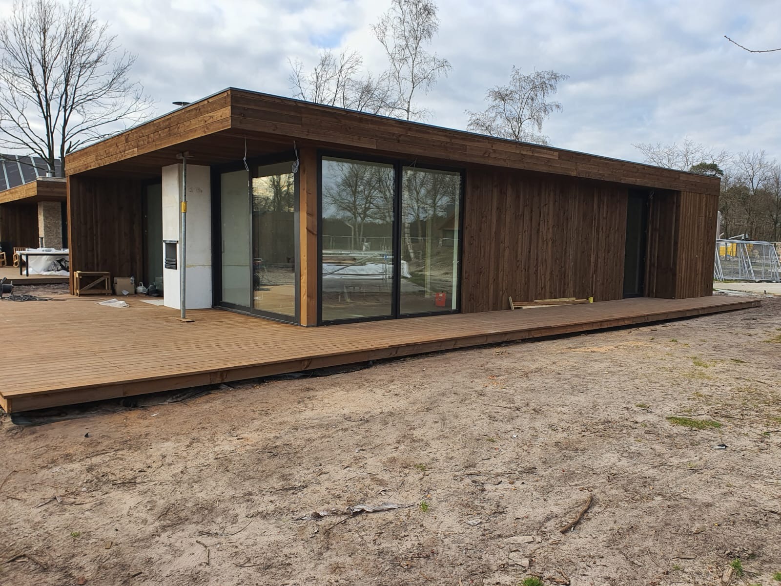Nowoczesny, parterowy budynek z drewnianą elewacją i dużymi przesuwnymi oknami, otoczony drewnianym tarasem. Widoczne drzewa w tle.