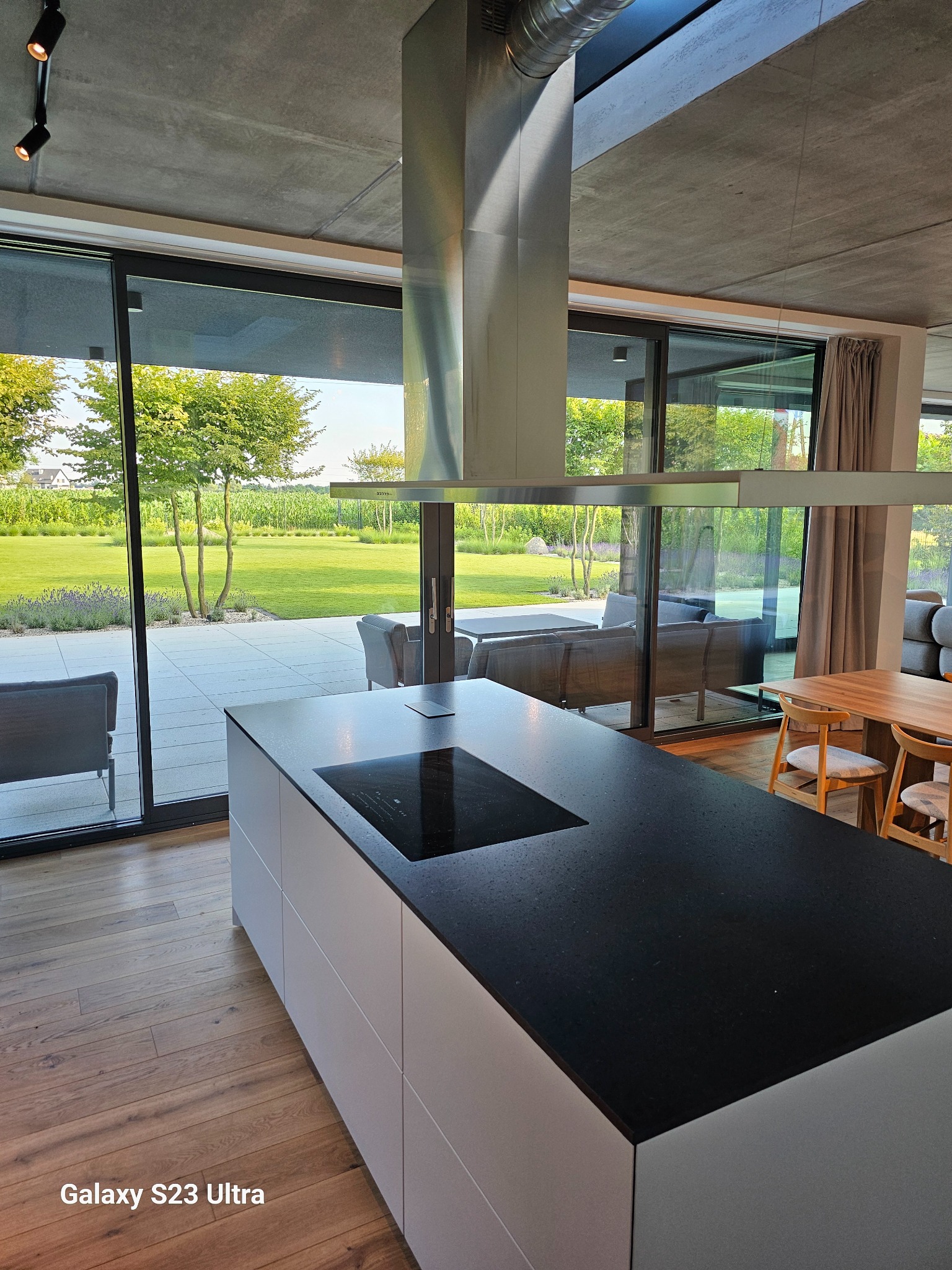 Nowoczesna wyspa kuchenna z czarnym blatem i białymi frontami na tle panoramicznego okna z widokiem na zielony ogród. Minimalistyczny design z metalowym okapem.