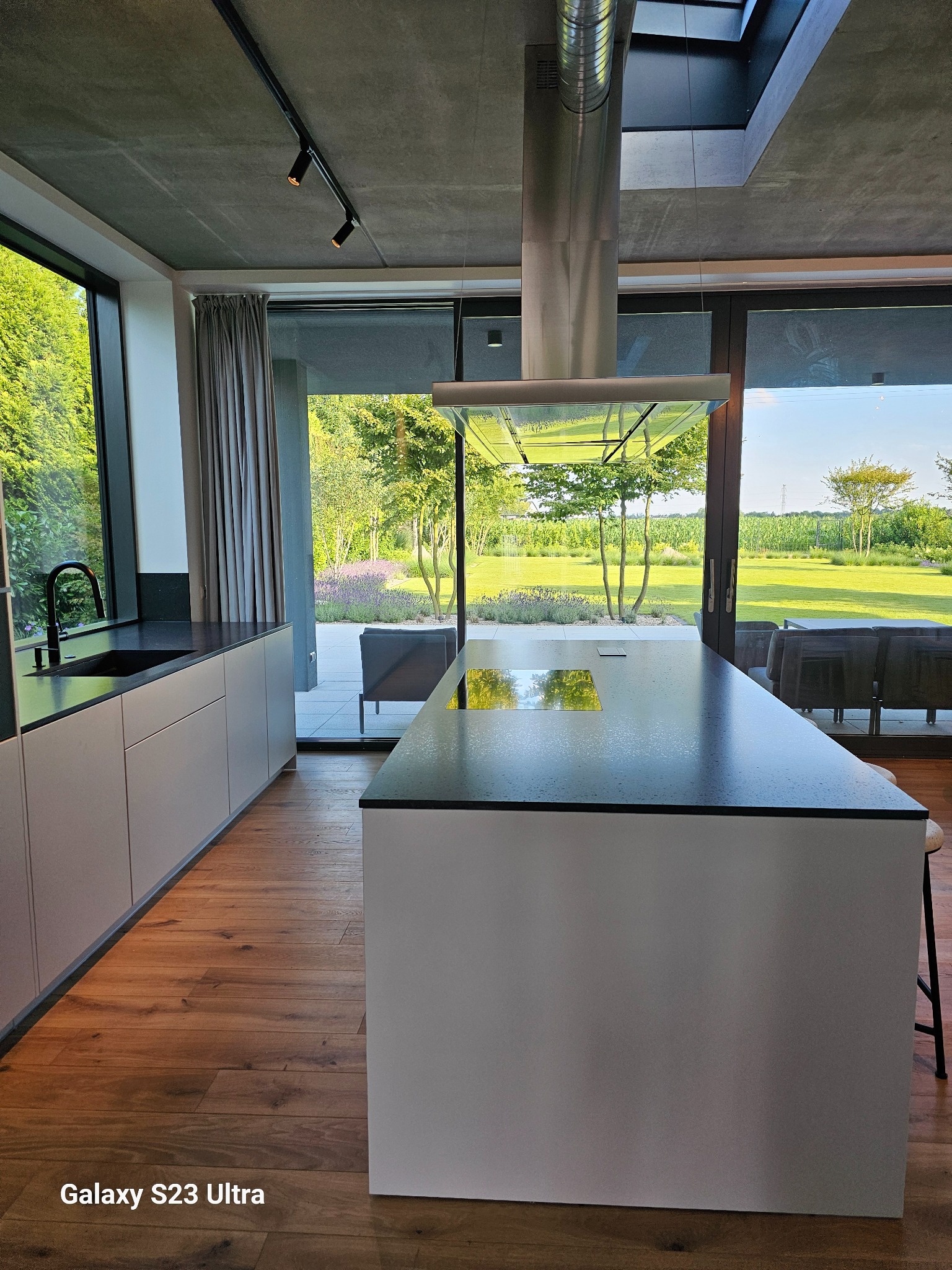 Nowoczesna kuchnia z wyspą i widokiem na ogród przez duże okna. Minimalistyczny design, drewniana podłoga, betonowy sufit z oświetleniem punktowym.