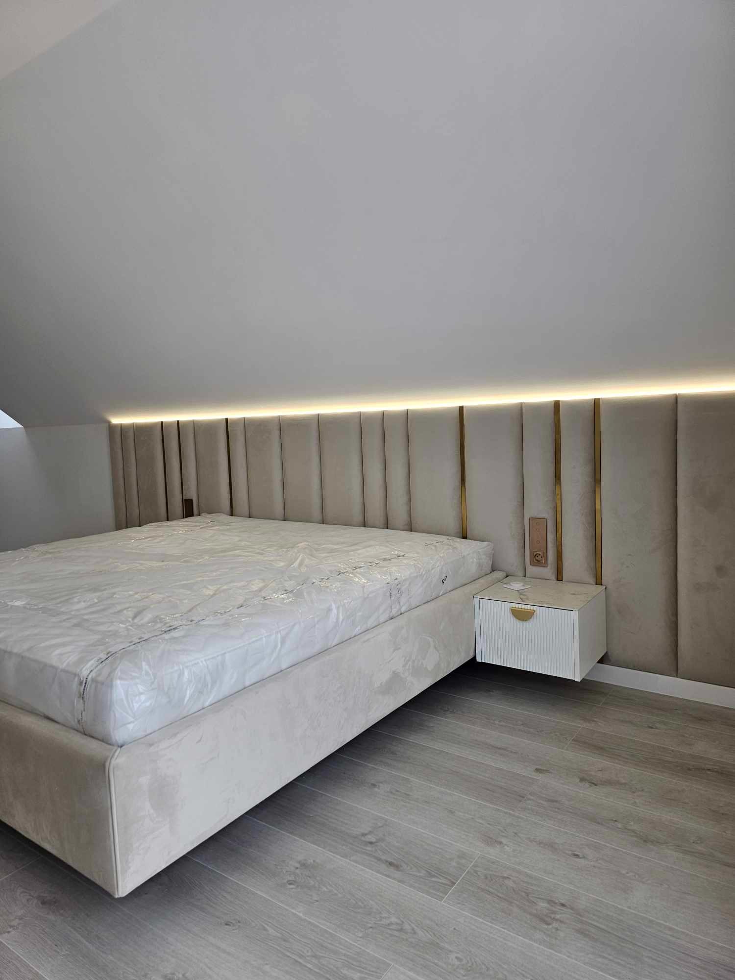 Beżowe łóżko z tapicerowanym wezgłowiem z oświetleniem LED i złotymi akcentami, szafka nocna w stylu glamour, szare panele podłogowe, pokój na poddaszu.