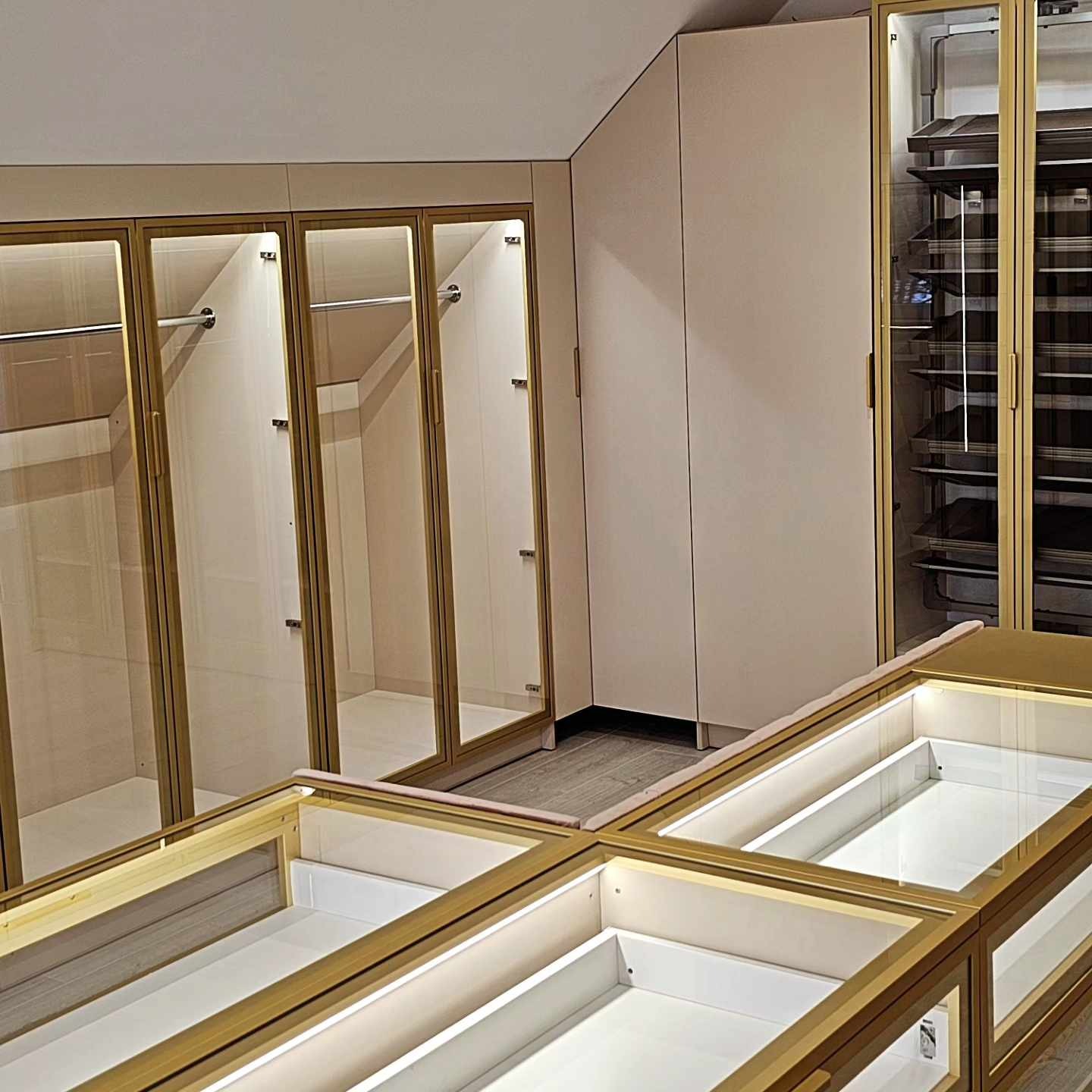 Nowoczesna garderoba z przeszklonymi drzwiami i złotymi ramami, oświetlone półki, beżowe ściany, widok z bliska na szuflady z oświetleniem LED.