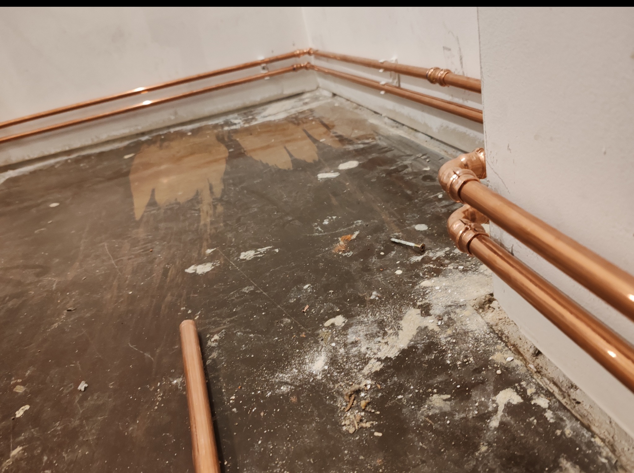Miedziane rury instalacji grzewczej zamontowane wzdłuż ścian, na podłodze widoczne ślady prac budowlanych. Rury łączone za pomocą złączek.