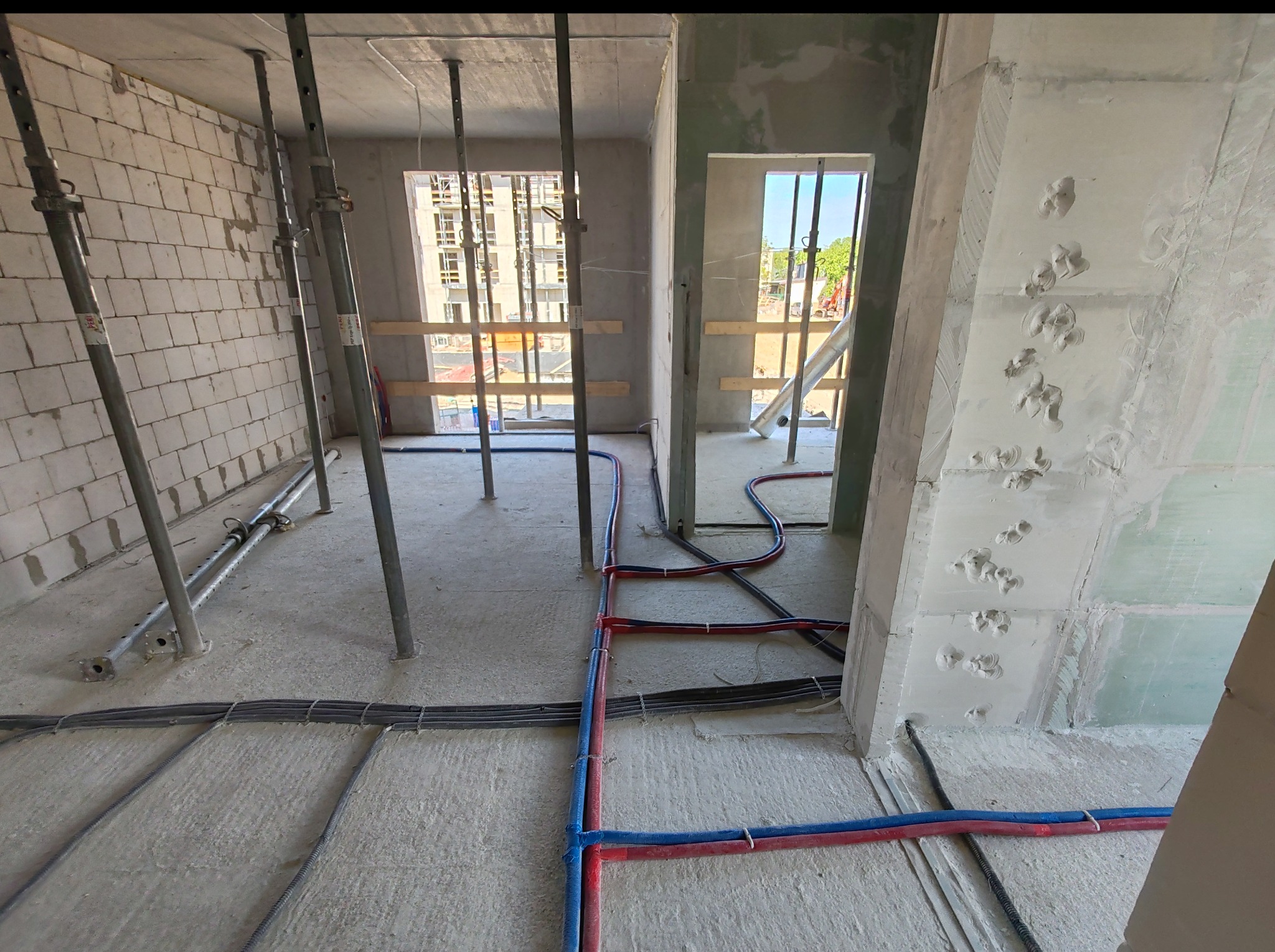 Surowy stan: instalacja wodna (czerwone i niebieskie rury) i elektryczna w peszlach ułożona na podłodze, ściany w tynku, podpory stropowe w tle okien.