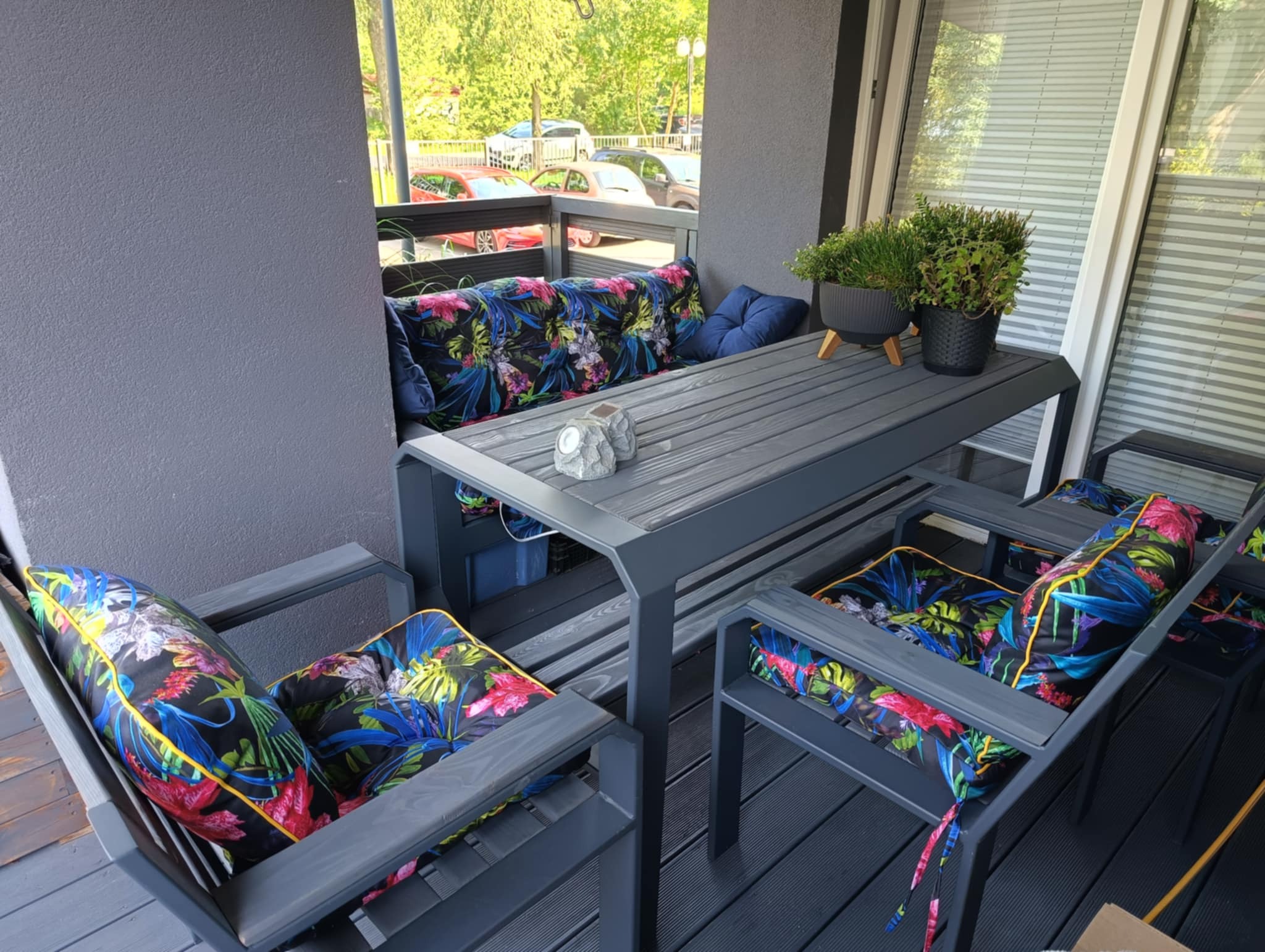 Szary zestaw mebli ogrodowych z poduszkami w tropikalne wzory na balkonie z drewnianą podłogą. Na stole doniczki z ziołami i dekoracje.