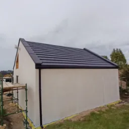 Nowy, grafitowy dach z blachy trapezowej na budynku z jasną elewacją. Widoczne obróbki blacharskie i rynny w kolorze dachu. Budynek w trakcie prac wykończeniowych, widoczne rusztowania.