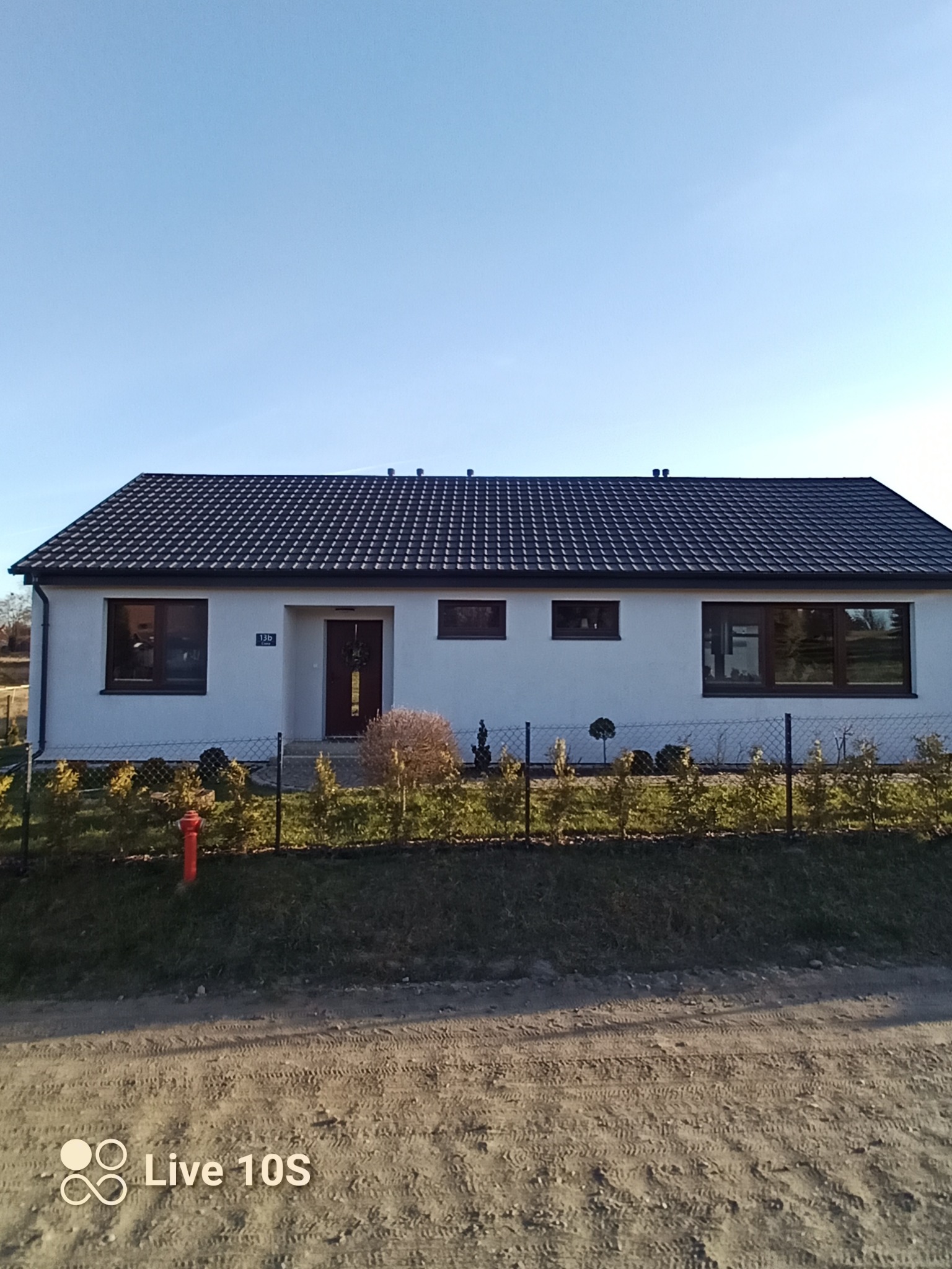 Jednopiętrowy dom z ciemną dachówką, białymi ścianami i brązowymi ramami okiennymi. Widoczny ogród z niskim żywopłotem i ścieżką. Czyste, błękitne niebo w tle.