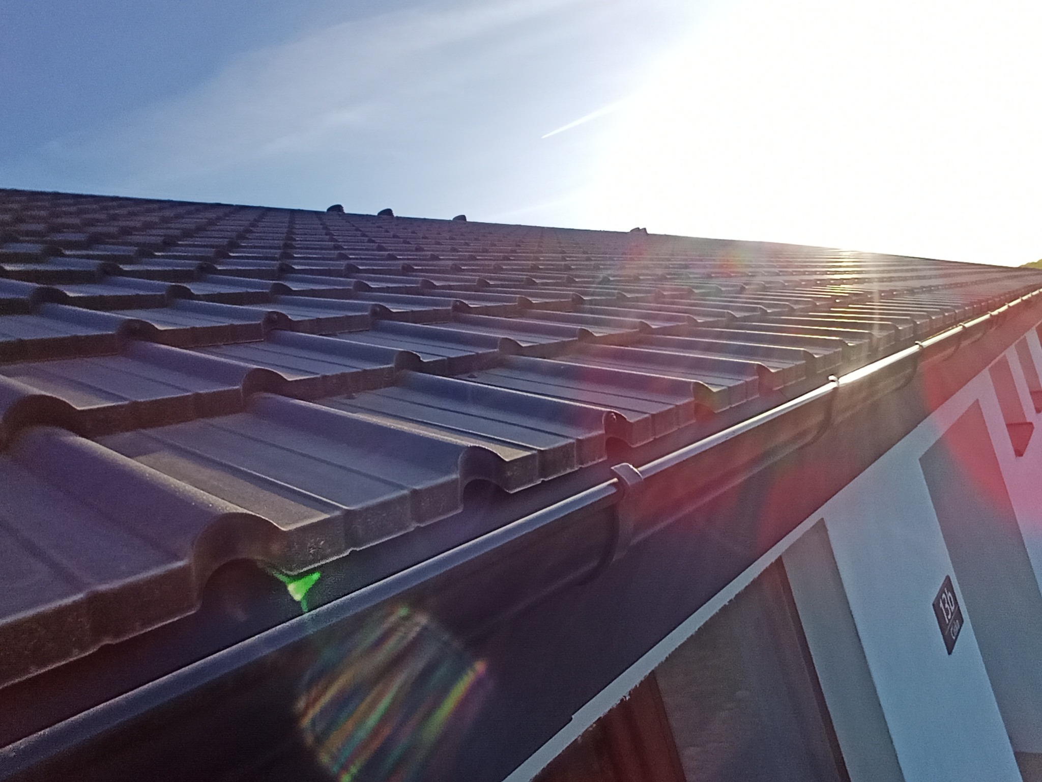 Nowy dach z ciemnej dachówki ceramicznej i czarną rynną, uchwycony pod kątem, z widocznym numerem domu 13b i fragmentem nieba z smugą kondensacyjną.