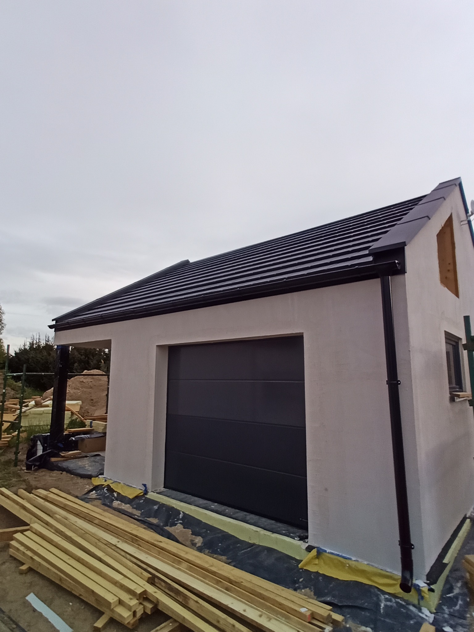 Nowoczesny garaż z czarnym dachem z dachówki i orynnowaniem. Widoczne deski i fragment okna. Budynek w trakcie wykończenia.