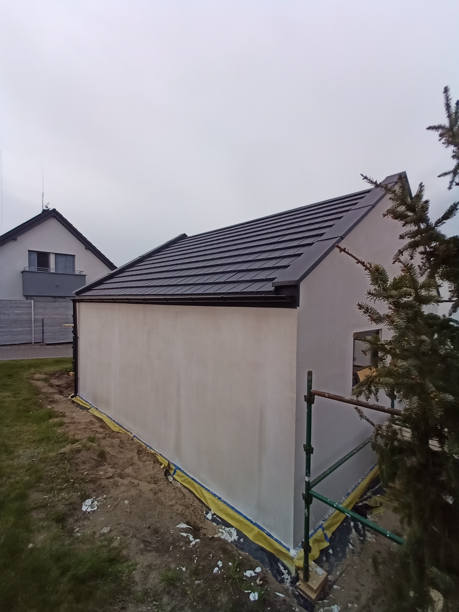 Nowy, grafitowy dach z blachodachówki na budynku z elewacją w trakcie prac. Widoczne rusztowanie i zabezpieczenia. W tle dom jednorodzinny i pochmurne niebo.