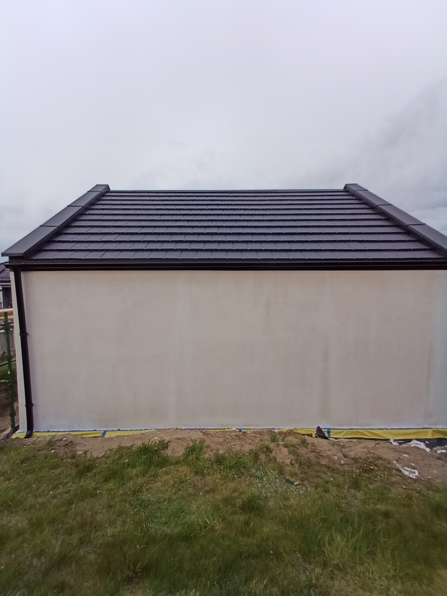 Ciemna dachówka ceramiczna na nowym dachu z zamontowanymi rynnami. Widok na fragment ściany budynku i teren wokół. Realizacja dekarska w pochmurny dzień.