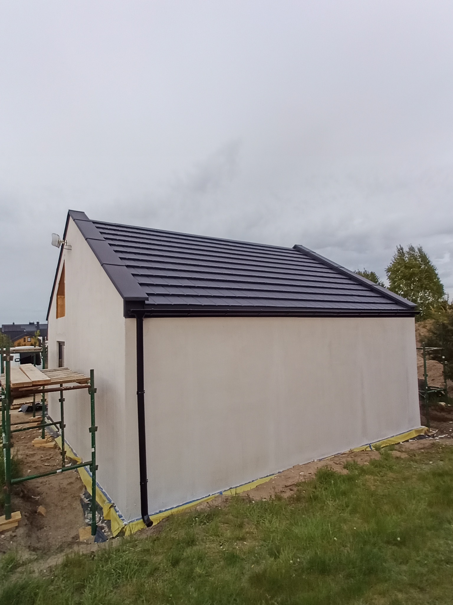 Nowy, grafitowy dach z blachy trapezowej na budynku z jasną elewacją. Widoczne obróbki blacharskie i rynny w kolorze dachu. Budynek w trakcie prac wykończeniowych, widoczne rusztowania.