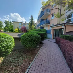 Ścieżka z kostki prowadząca do wejścia budynku z balkonami, otoczona równo przystrzyżonymi krzewami i drzewami w słoneczny dzień.