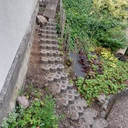 LDN PERFECT - Ułożone schody z ażurowych betonowych płyt na zboczu, z widoczną ziemią i roślinnością. Drewniane paliki wzdłuż ścieżki.