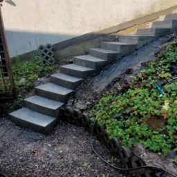 Betonowe schody prowadzące w górę zbocza, otoczone roślinnością i kamieniami. Ściana w tle z oświetleniem. Widok z dołu.