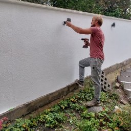 LDN PERFECT - Instalacja oświetlenia zewnętrznego: mężczyzna montuje lampę na białym murze, używając wkrętarki. Widoczne rośliny u podnóża muru i drzewa w tle.