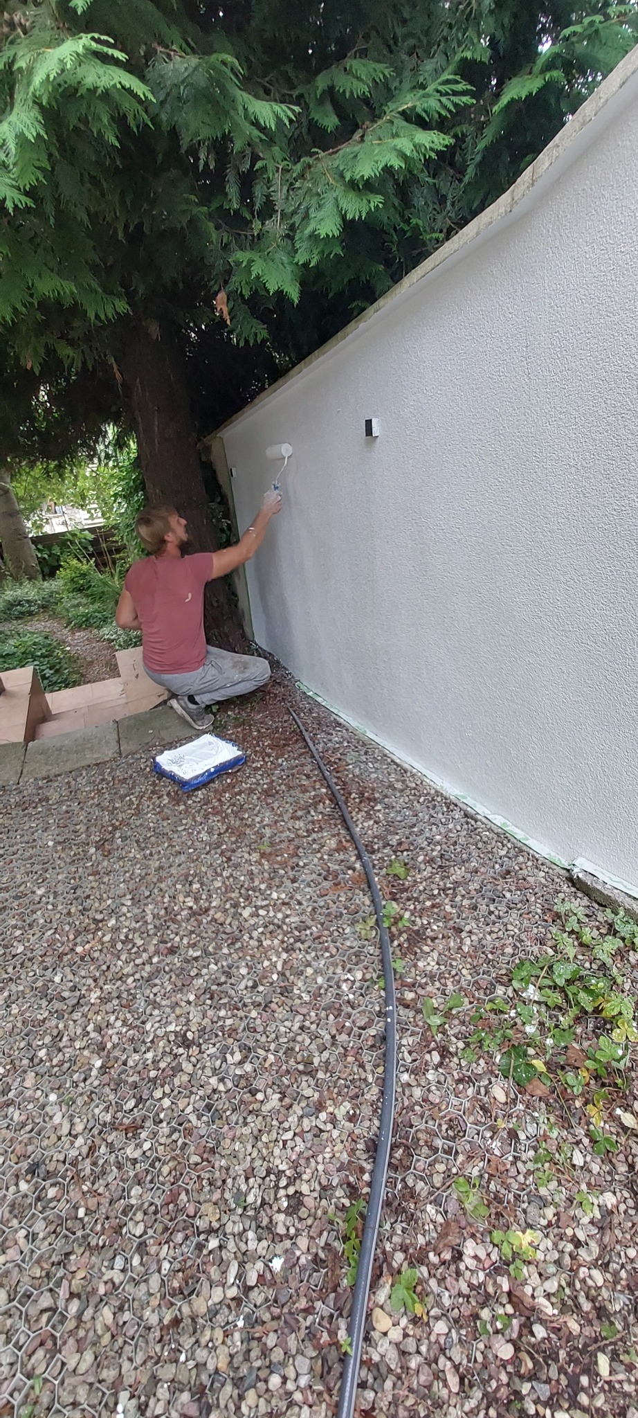 Malowanie elewacji: mężczyzna w trakcie pracy, siedzi na kamiennym podłożu i wałkiem nanosi białą farbę na szorstką ścianę. Obok drzewo.