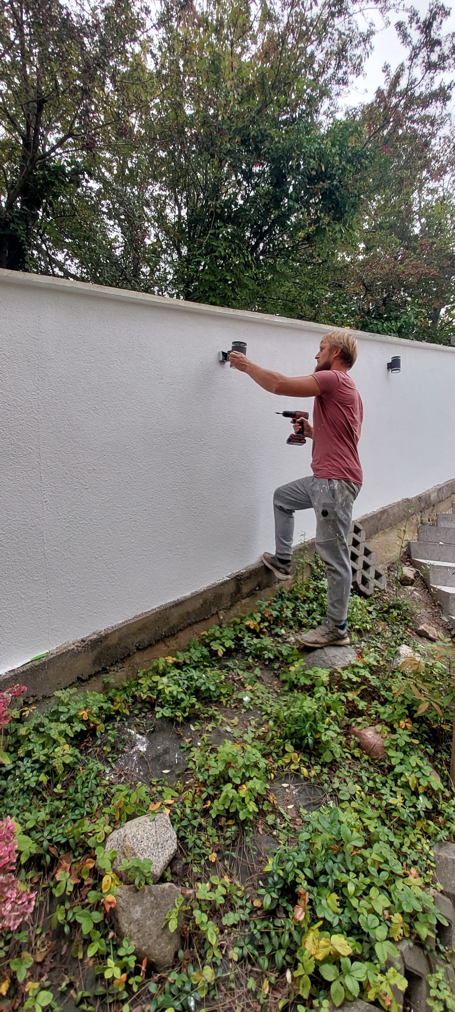 Instalacja oświetlenia zewnętrznego: mężczyzna montuje lampę na białym murze, używając wkrętarki. Widoczne rośliny u podnóża muru i drzewa w tle.