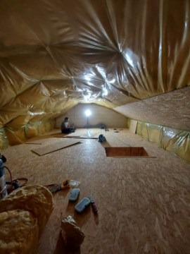 Adaptacja poddasza: izolacja folią i wykończenie płytami OSB. Widoczny otwór w podłodze, narzędzia i pracownik w oddali. Oświetlenie punktowe.