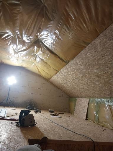 Poddasze w trakcie izolacji: folia paroizolacyjna na skośnym suficie, płyty OSB na ścianie, widoczna piła tarczowa i oświetlenie robocze. Prace wykończeniowe.