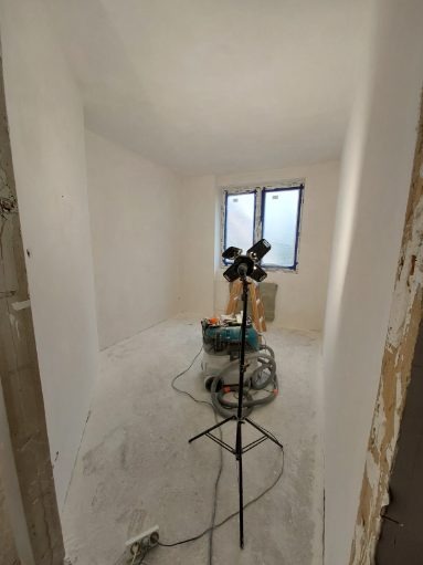 Pokój po gładzi, przygotowany do malowania. Widoczne narzędzia: odkurzacz przemysłowy, lampa na statywie. Białe ściany, okno z niebieską ramą.