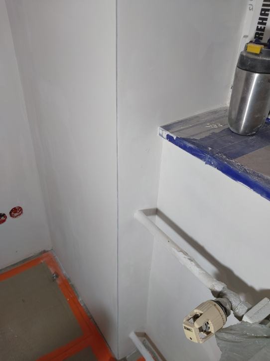 Gładka, biała ściana po malowaniu, widoczne elementy instalacji grzewczej i zabezpieczona folią półka. Detal wykończenia wnętrza z bliska.