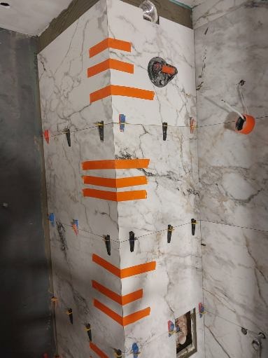 Układanie marmurowych płytek w rogu łazienki, z użyciem pomarańczowych pasków i czarnych klipsów poziomujących, widoczny fragment instalacji wodnej.