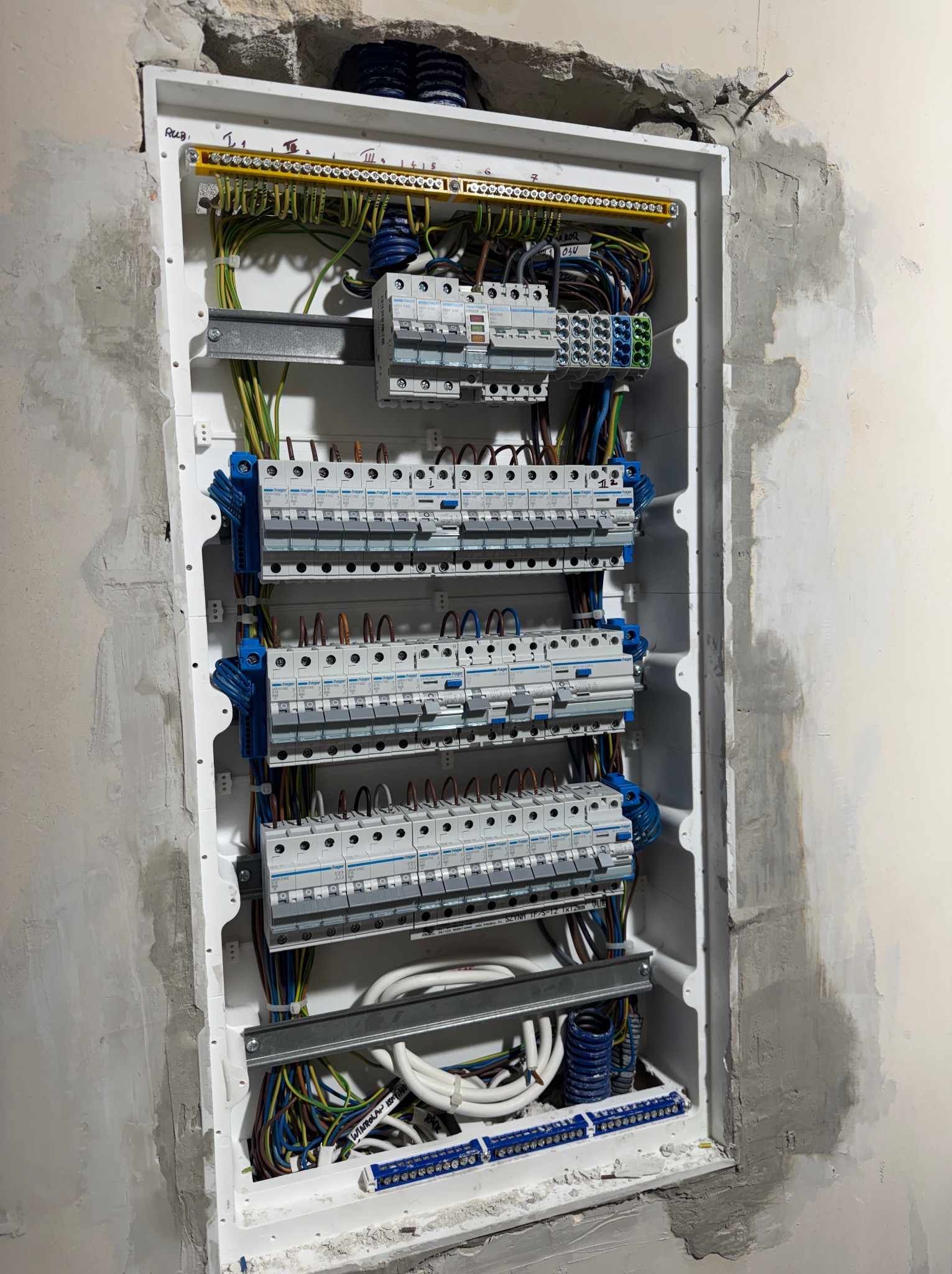 Rozdzielnica elektryczna Hager w trakcie montażu w ścianie, widoczne przewody, bezpieczniki i listwy zaciskowe. Ściana w stanie surowym, przygotowana do tynkowania.