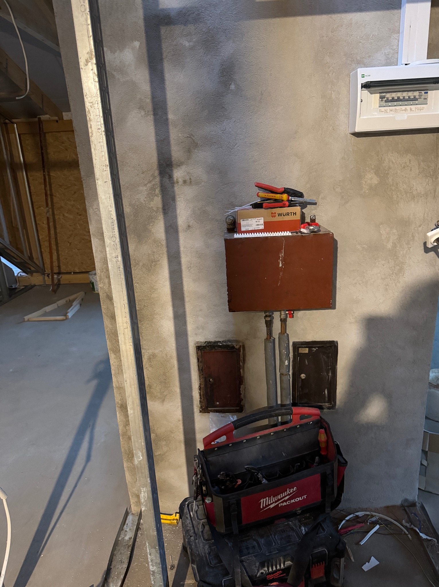 Szara ściana z brązową skrzynką, narzędzia Wurth, skrzynka Milwaukee Packout. Widoczna instalacja i skrzynka z bezpiecznikami. Surowy stan wykończenia wnętrza.