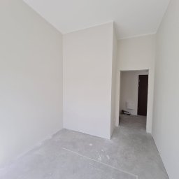 SPOKOJNA BUDOWA GVO SP. Z O.O. - Jasne, świeżo pomalowane wnętrze pokoju z szarym betonowym podłożem i widocznym wejściem do kolejnego pomieszczenia. Minimalistyczny design, neutralna kolorystyka.