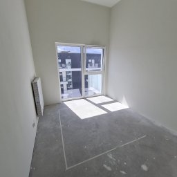 SPOKOJNA BUDOWA GVO SP. Z O.O. - Wnętrze pokoju w stanie deweloperskim z oknem i świeżo pomalowanymi ścianami na biało. Na podłodze widoczne ślady po pracach wykończeniowych.