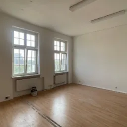 Jasne, puste pomieszczenie z dwoma oknami, drewnianą podłogą i świeżo pomalowanymi ścianami. Na podłodze widoczne puszki z farbą, sugerujące etap prac wykończeniowych.