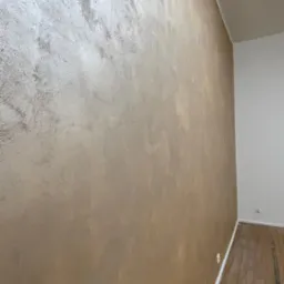 Ściana z efektem dekoracyjnym w odcieniach beżu i srebra, widoczna pod kątem w jasnym wnętrzu. Na podłodze parkiet, a w rogu biała ściana.