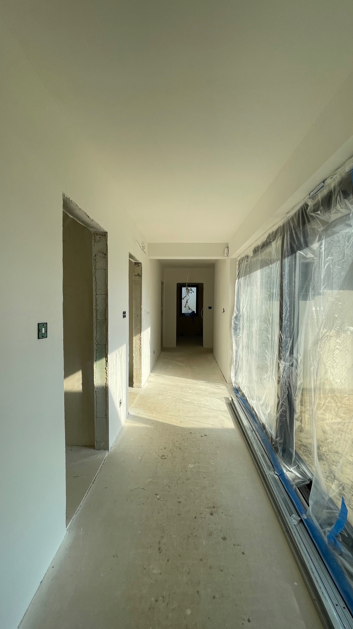 Korytarz w trakcie wykańczania: świeżo pomalowane ściany, surowa posadzka, okna zabezpieczone folią. Widoczne ślady szpachlowania i przygotowania do dalszych prac.