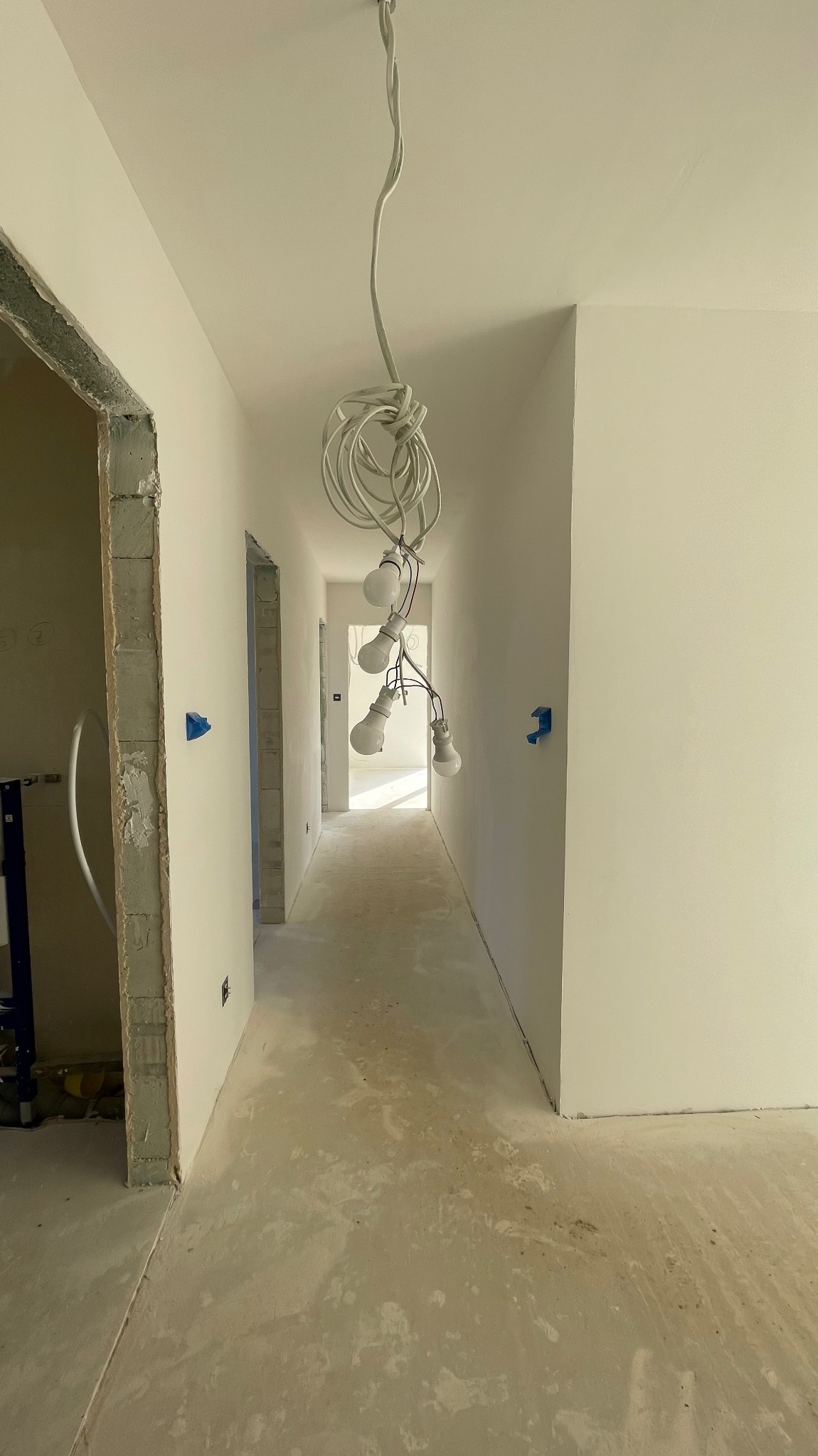 Korytarz w trakcie prac wykończeniowych: świeżo pomalowane ściany na biało, surowa podłoga, tymczasowe oświetlenie wiszące. Widoczne fragmenty instalacji.