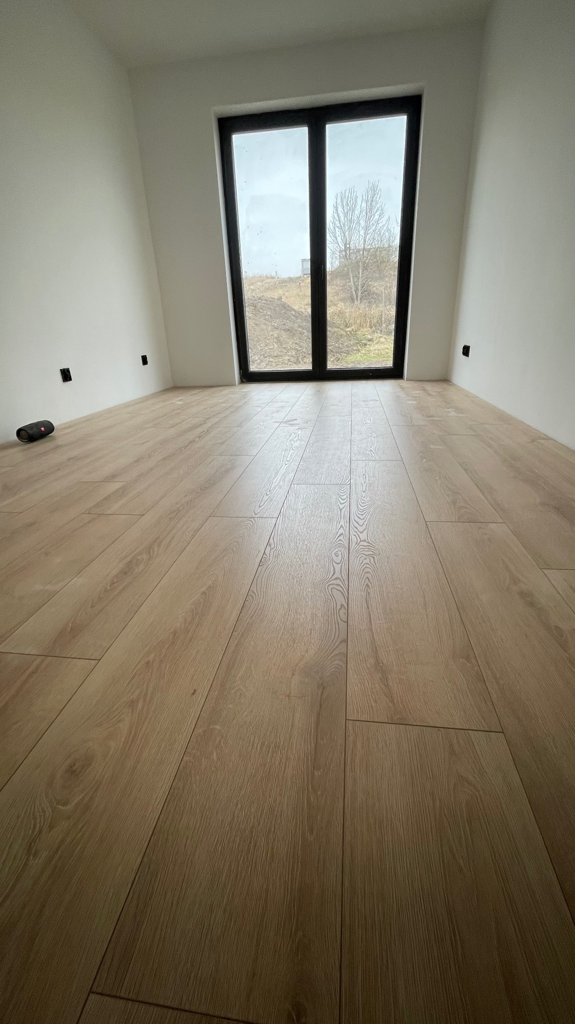 Jasna podłoga panelowa w nowym pokoju z oknem na zewnątrz. Widoczne gładkie, białe ściany i czarne gniazdka elektryczne. Minimalistyczny, nowoczesny styl.