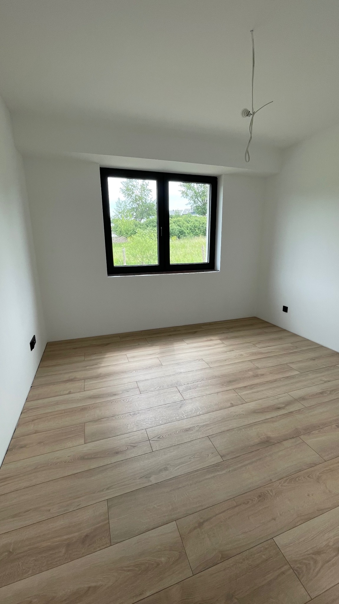 Jasne panele podłogowe w pokoju z białymi ścianami i oknem z widokiem na zieleń. Widoczne gniazdka elektryczne i wystające kable oświetleniowe.