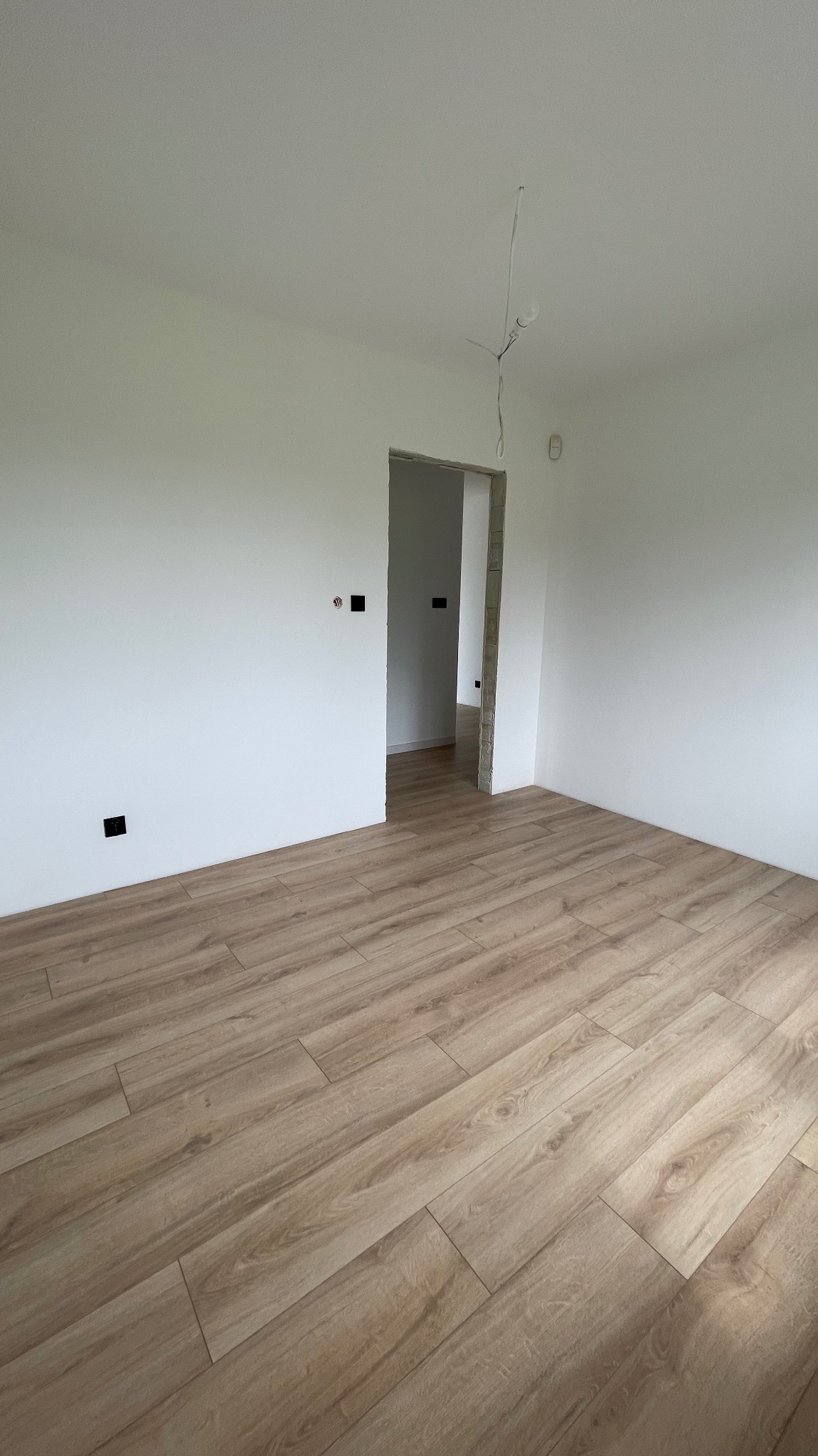 Jasne panele podłogowe ułożone w pustym pokoju z białymi ścianami i otwartym przejściem do kolejnego pomieszczenia, widoczne gniazdka elektryczne.